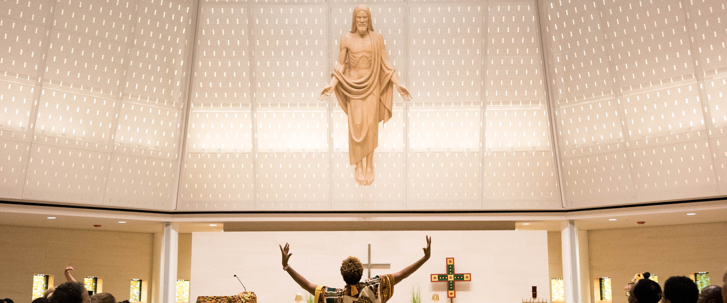 A church sanctuary with a large statue of Jesus suspended above the altar, arms open, while a worship leader below raises their arms in praise; a cross and congregants are visible in the background.