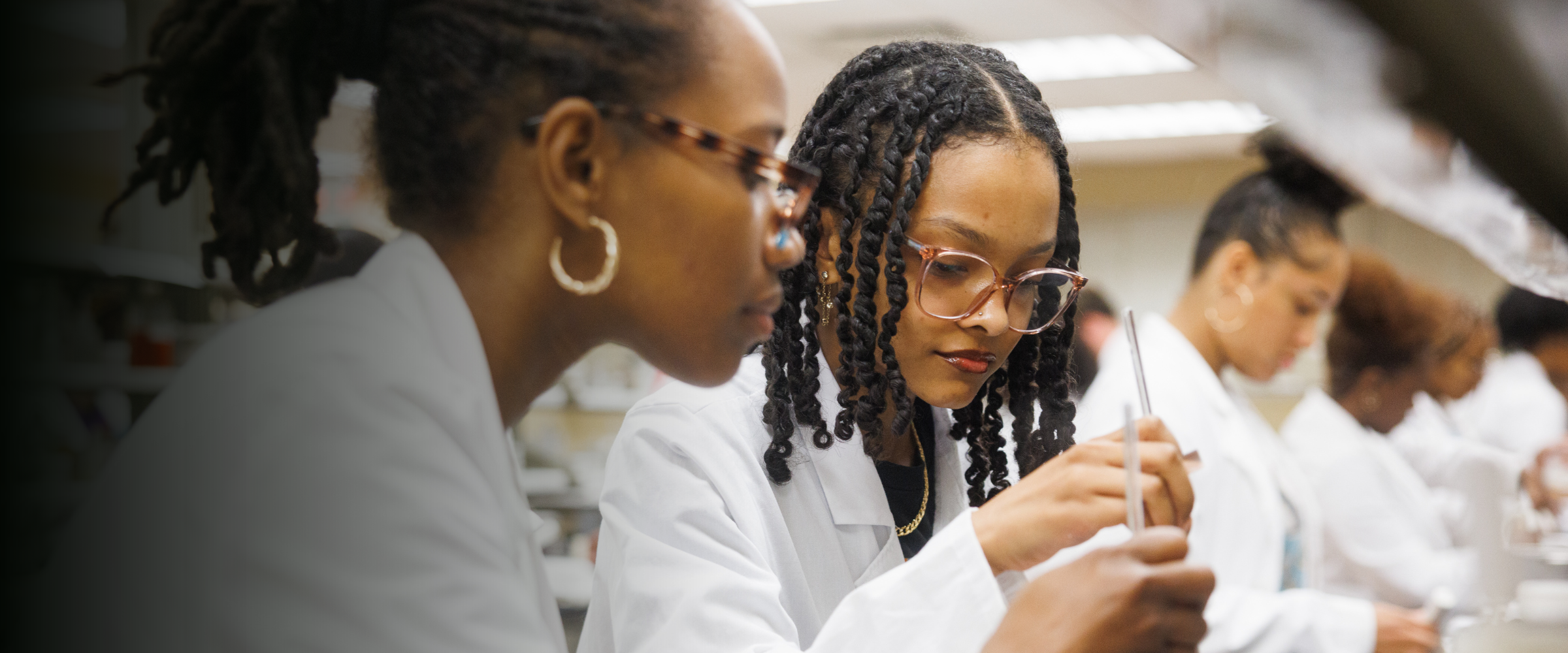 Two female pharmacy students wearing white coats mixing a solution (front) among other students in the laboratory (back)