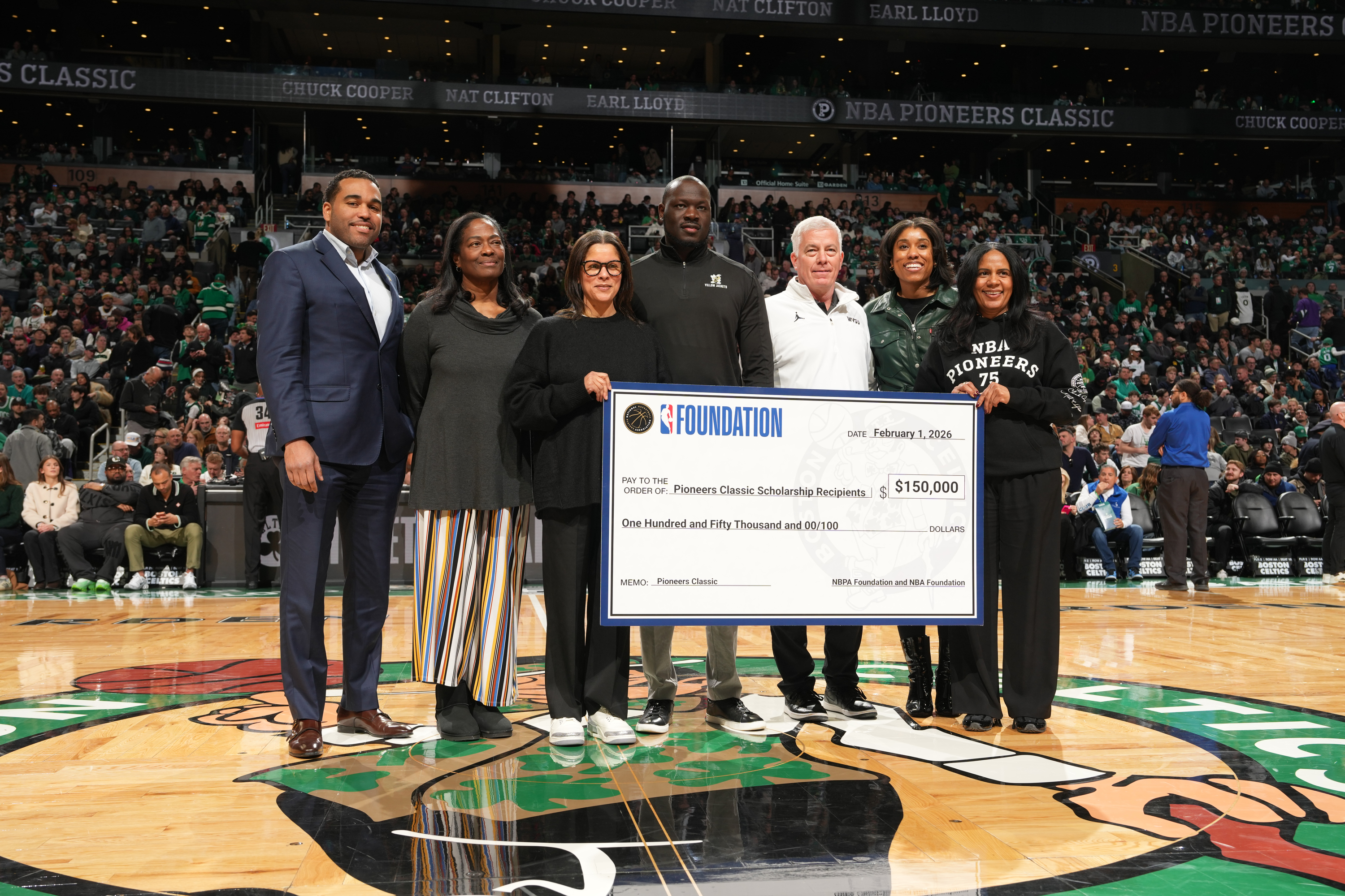 In a stadium full of people, seven people stand on a basketball court that sports the Boston Celtics logo, holding a large $150,000 check made to the Pioneers Classic Scholarship Recipients. Among them stand Ronald Carrere Jr. (left) and Pat Kendrick (second to left).