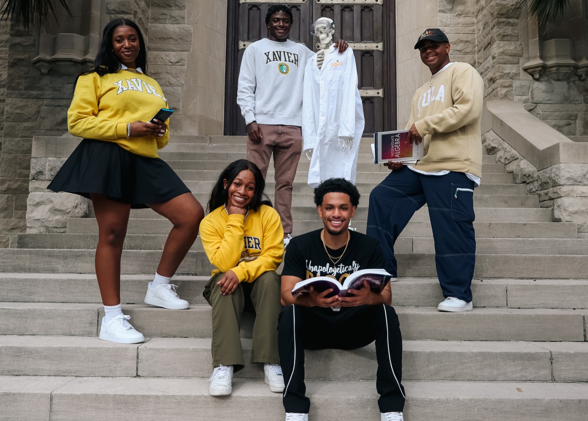 Five students standing outside on a staircase at Xavier University of Louisiana with a skeleton wearing a lab coat.
